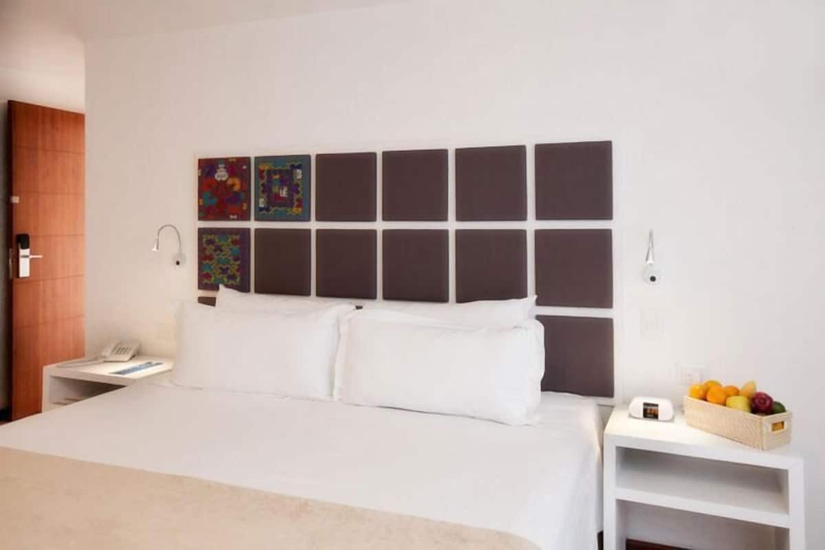 Habitación de hotel con una cama grande, una mesita de noche con una cesta de frutas y una pared con paneles cuadrados.