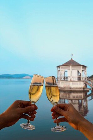 Two hands holding wine glasses with a calm body of water and a building in the background. - Hotel Butterfly - We Suite (Viareggio)