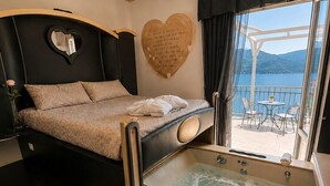 A bedroom with a bed, a heart-shaped wall decoration, a bathtub, and a balcony with a view of the water. - Hotel Butterfly - We Suite (Viareggio)