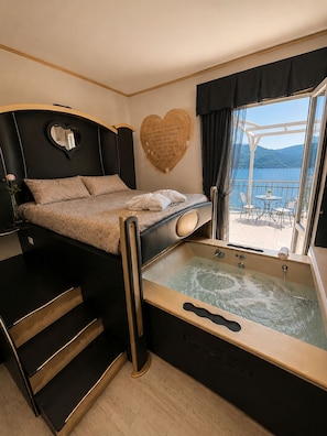 A bedroom with a bed, a heart-shaped wall decoration, a bathtub, and a balcony with a view of the water. - Hotel Butterfly - We Suite (Viareggio)