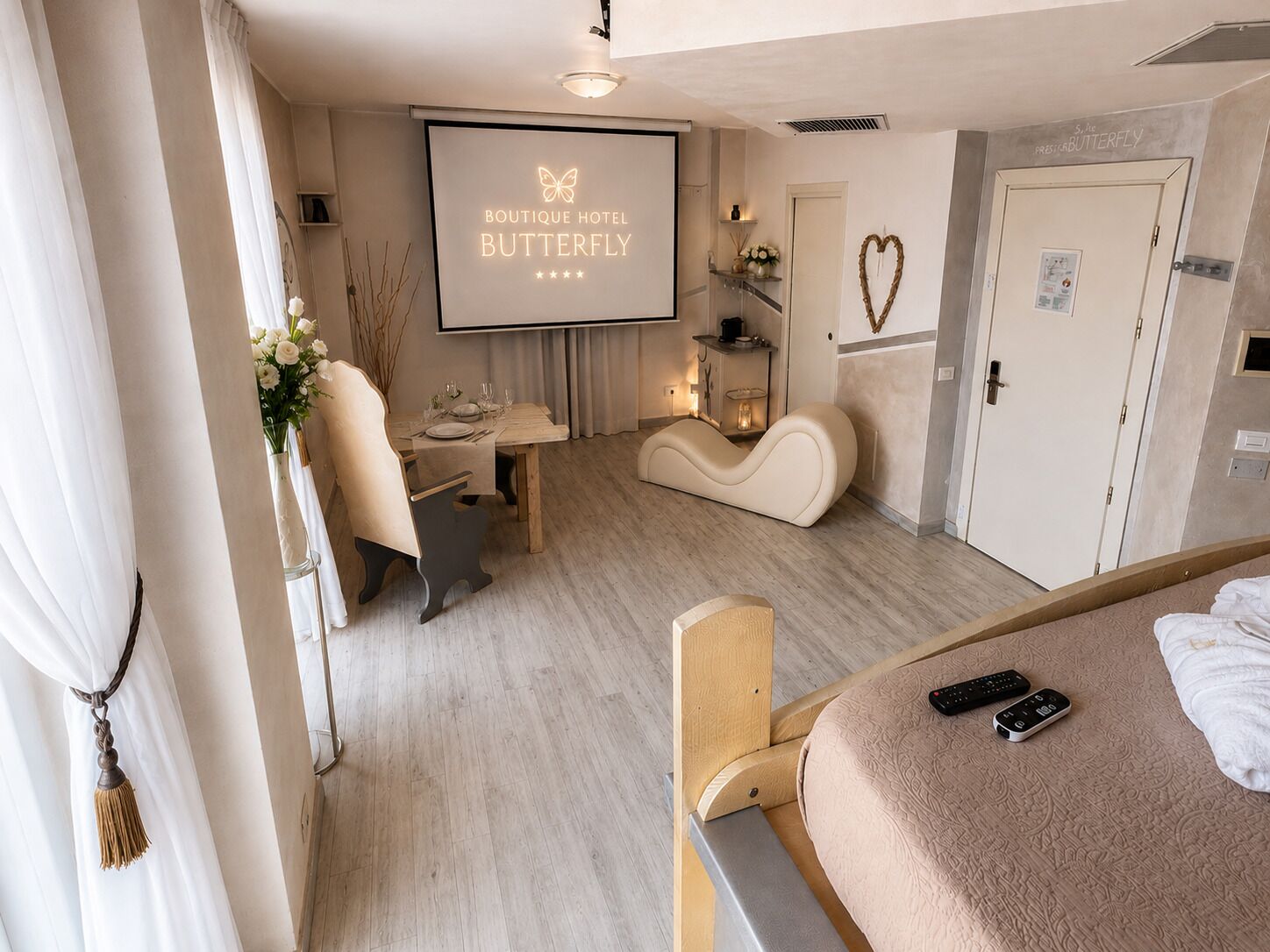 Ein Hotelzimmer mit einem Bett, einem Schreibtisch, einem Sessel, einem Fernseher, der den Hotelnamen anzeigt, und einer herzförmigen Dekoration an der Wand.