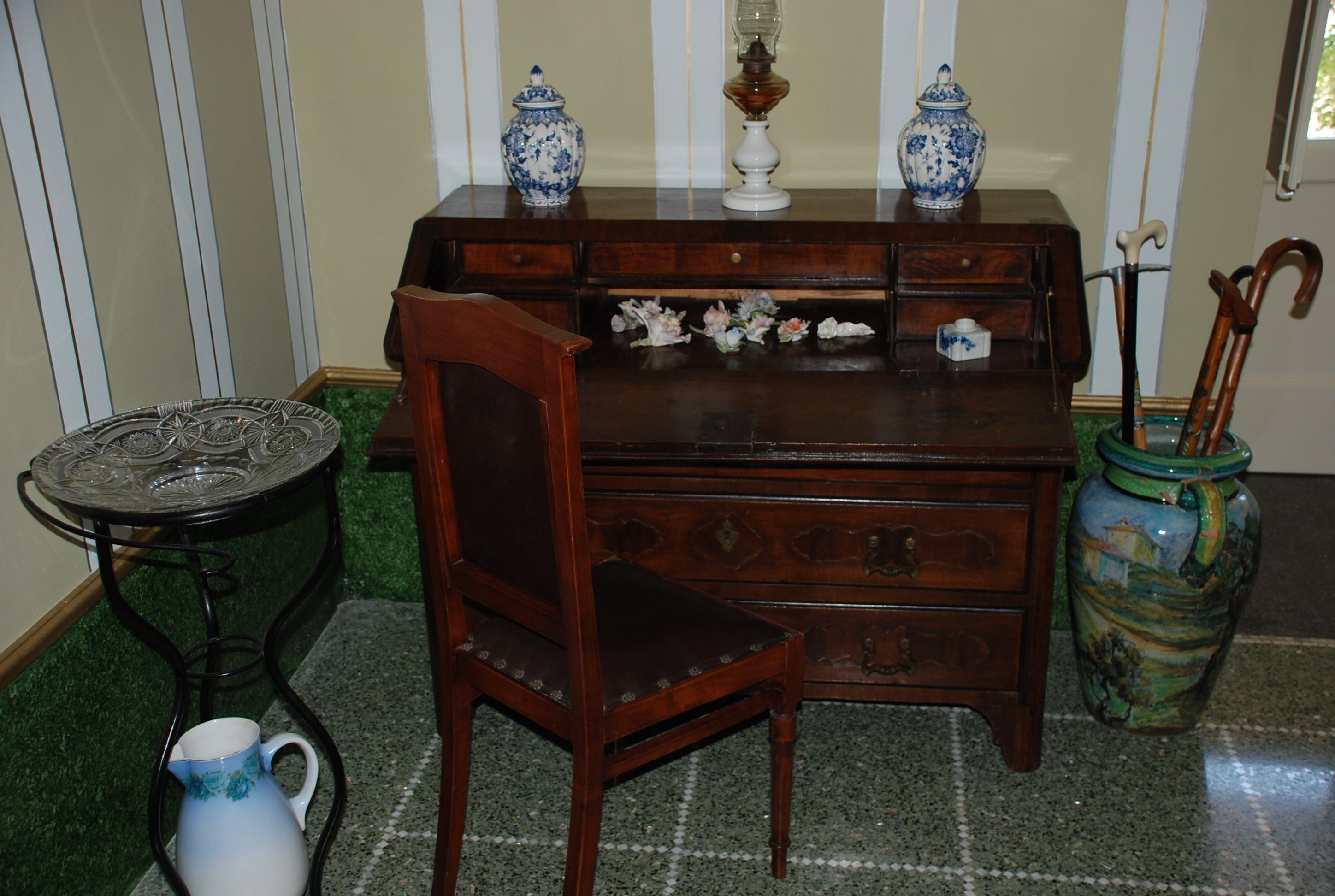 A wooden chest of drawers with floral patterns, a ceramic pitcher, and a vase with a cane.