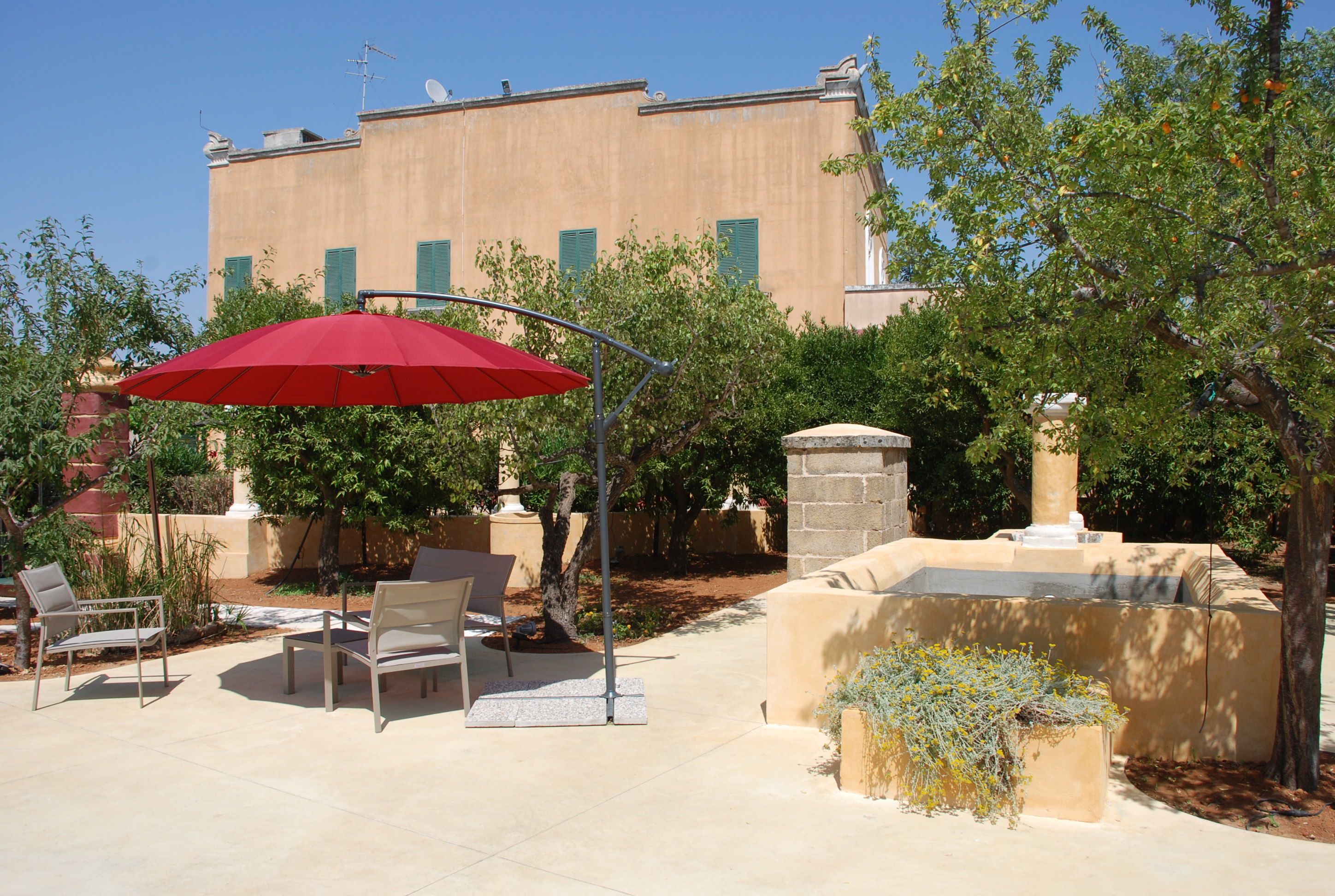 An outdoor patio with a red umbrella, two chairs, and a stone planter with plants.