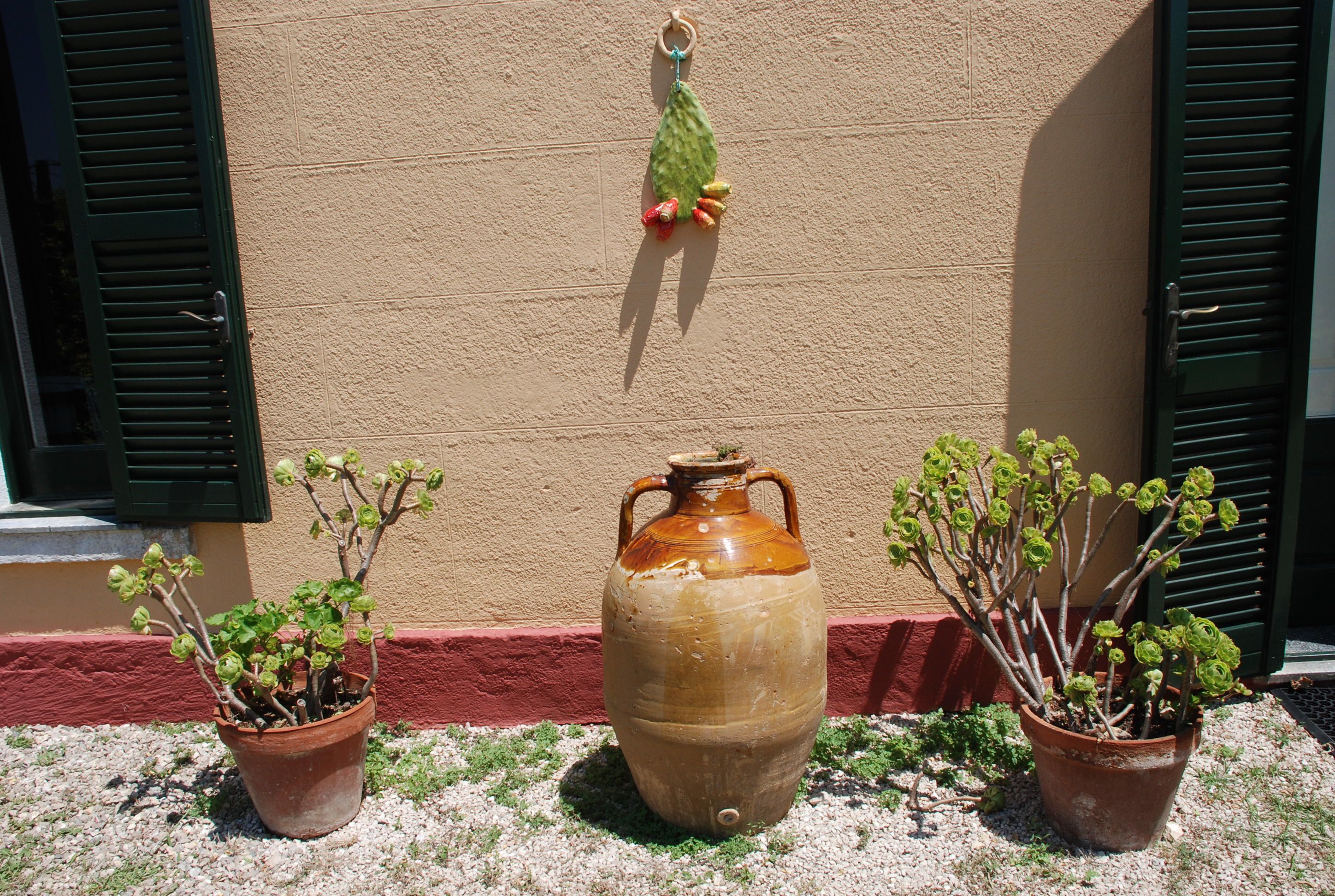 Een grote terracotta pot met handgrepen, twee kleinere terracotta potten met planten, en een muur met rode details en groene luiken.