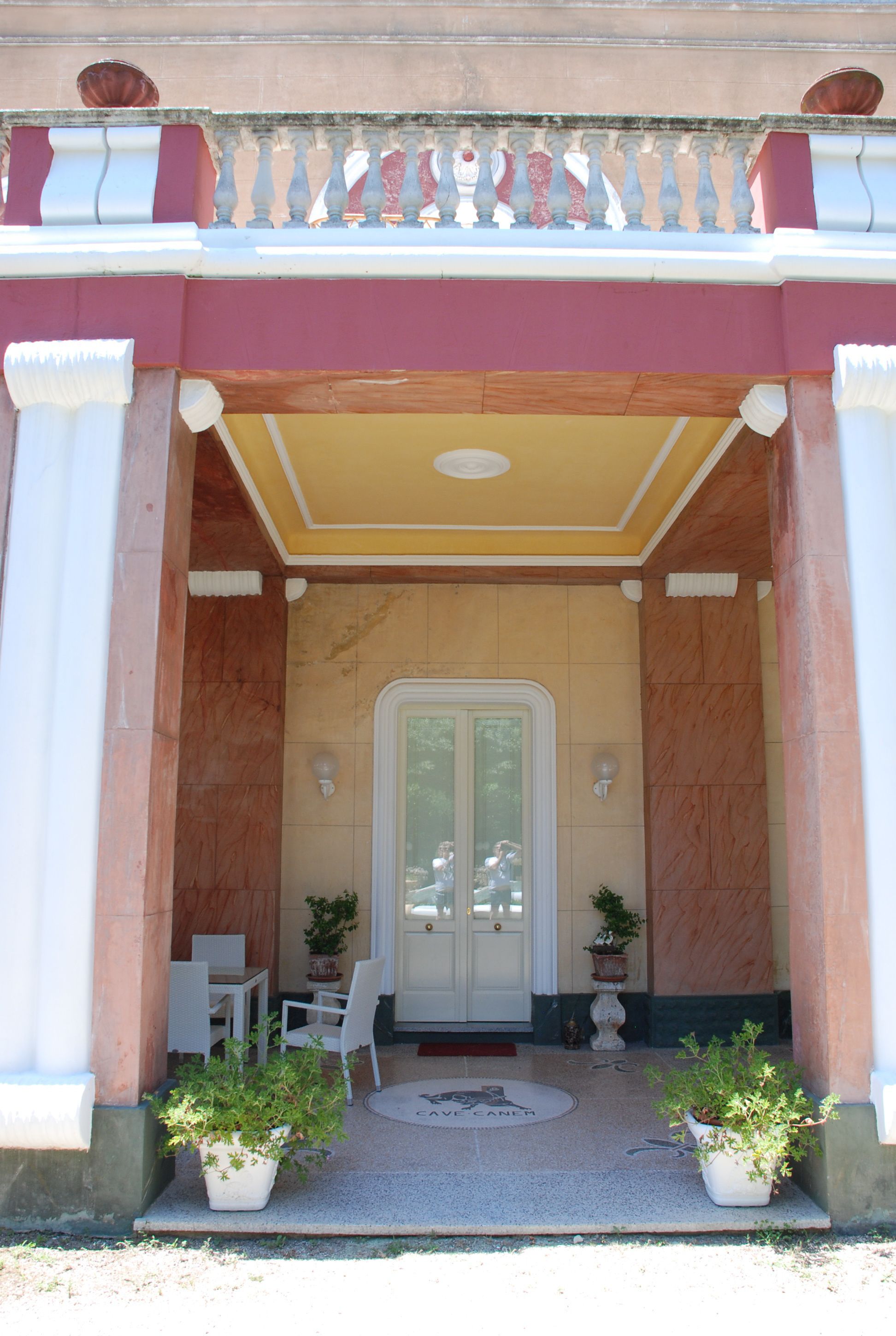 A building entrance with a yellow ceiling, white columns, and a door with glass panels. There are two potted plants on either side of the door.