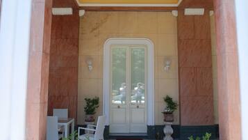 A building entrance with a yellow ceiling, white columns, and a door with glass panels. There are two potted plants on either side of the door.