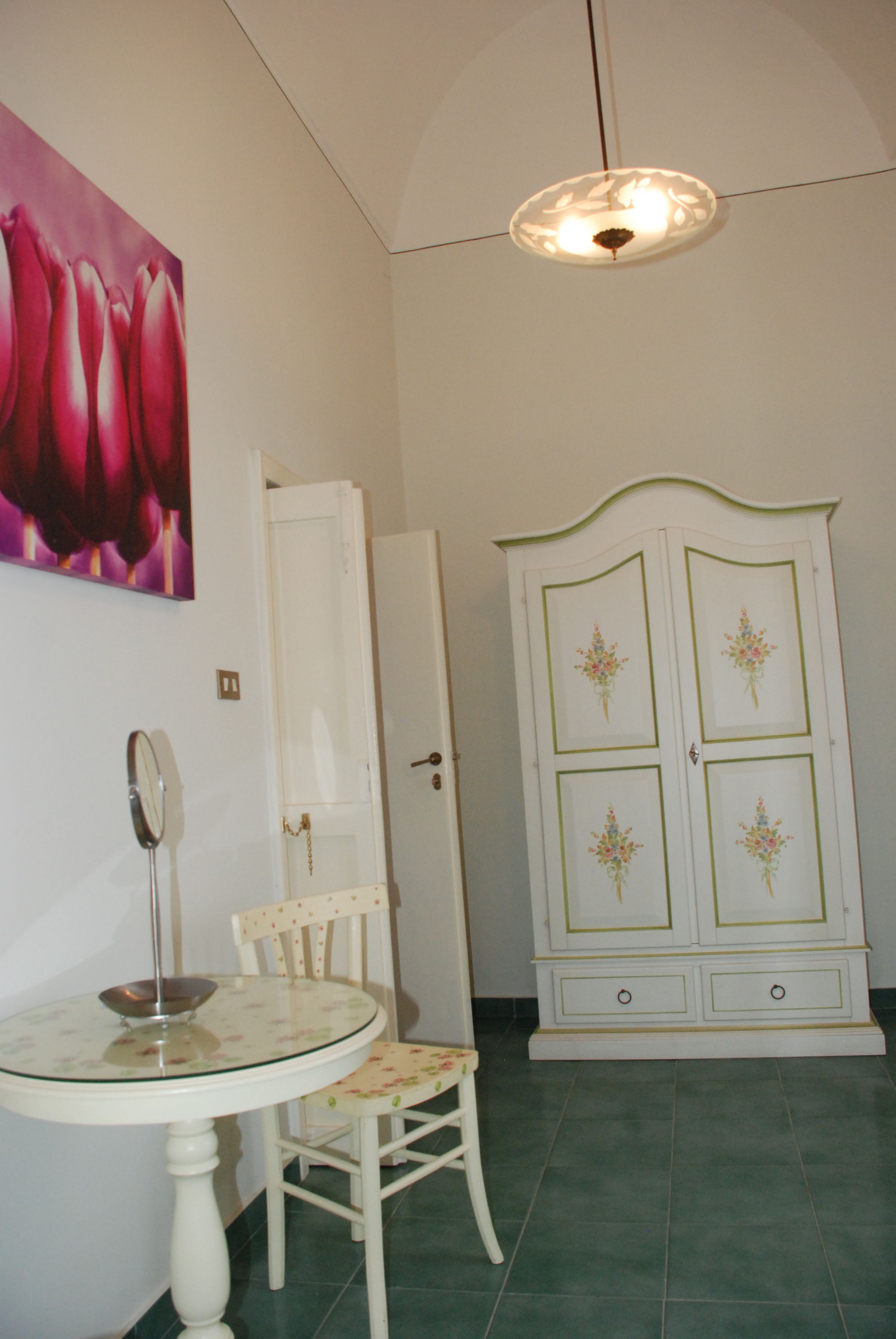 Une pièce avec une table ronde en verre, une chaise blanche, un meuble blanc orné de motifs floraux et un tableau représentant des fleurs accroché au mur.