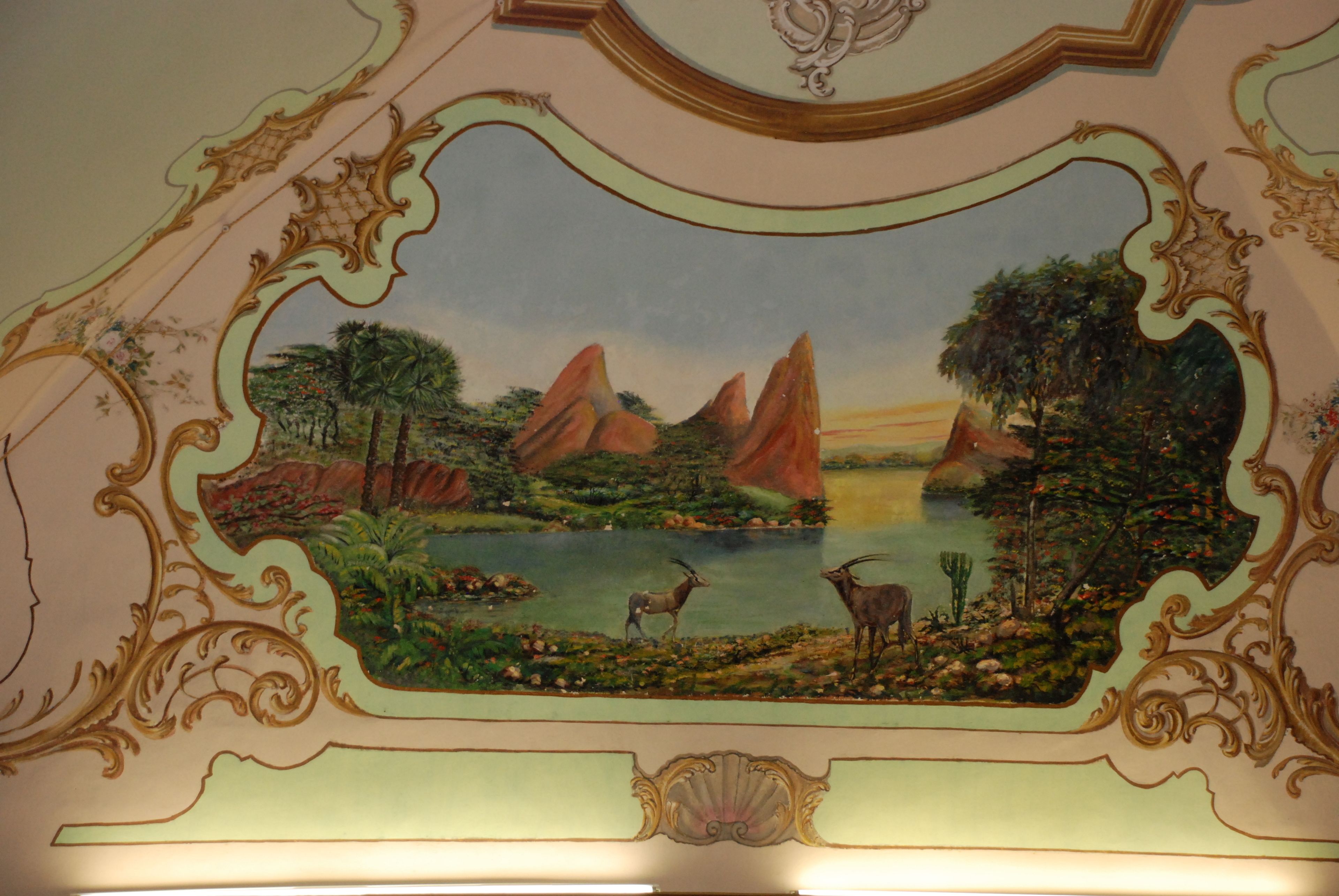 Un mural en el techo con una escena de paisaje, detalles ornamentales dorados y un elemento decorativo central.