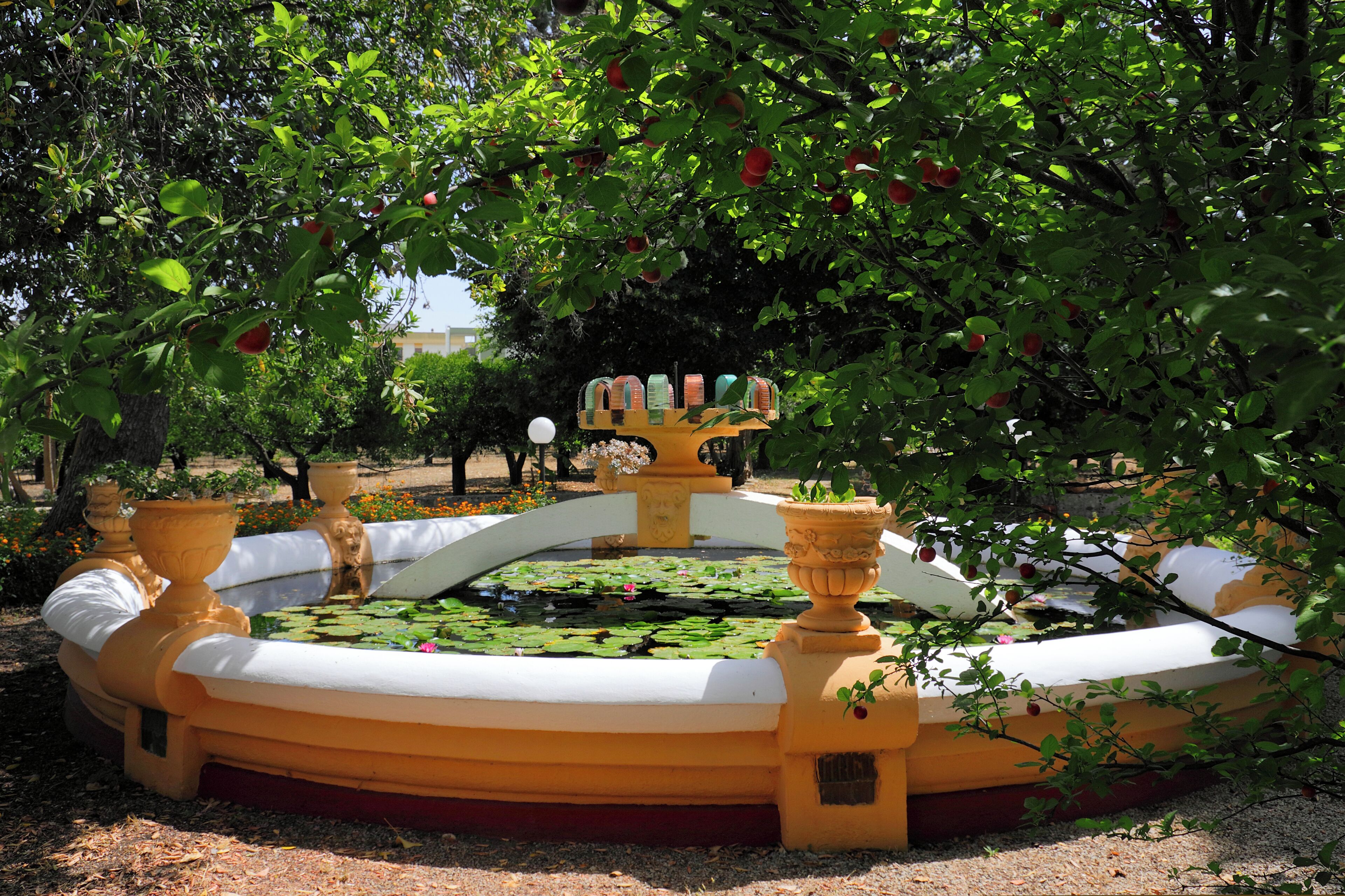 A circular fountain with a central statue, surrounded by a yellow and white tiled area, with trees and flowers in the background.