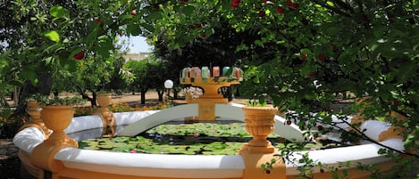 A circular fountain with a central statue, surrounded by a yellow and white tiled area, with trees and flowers in the background.