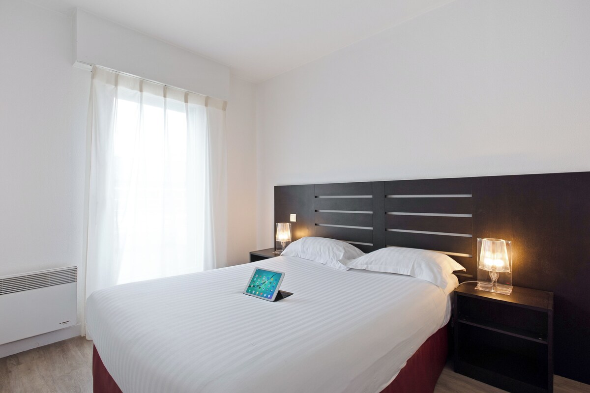 A hotel room with a large bed, a bedside table with a lamp, a tablet on the bed, and a window with curtains.