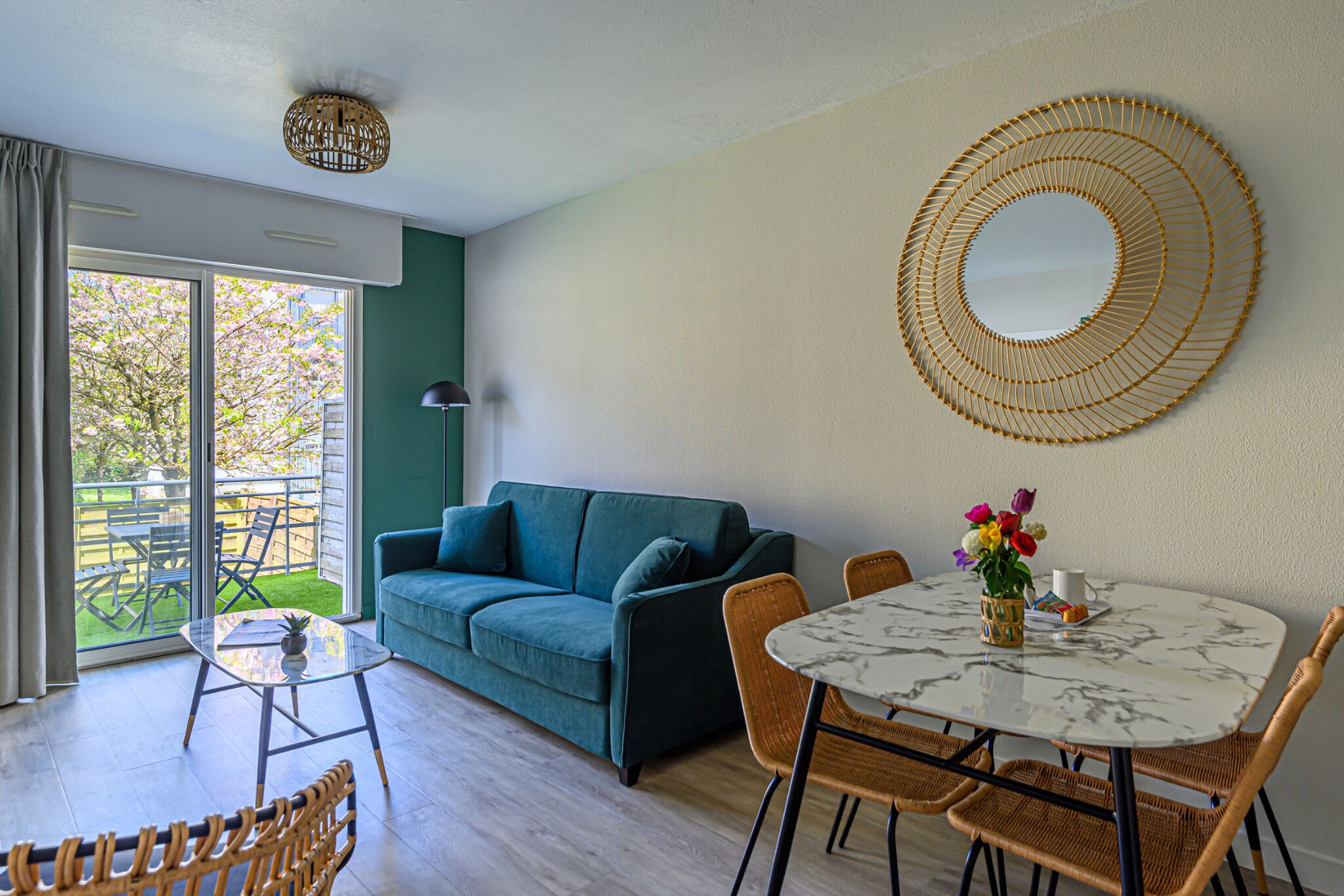 Ein modernes Wohnzimmer mit einem grünen Sofa, einem runden Tisch mit Stühlen, einem großen Spiegel und Blick auf einen Garten durch eine Schiebetür.