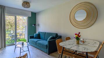 Ein modernes Wohnzimmer mit einem grünen Sofa, einem runden Tisch mit Stühlen, einem großen Spiegel und Blick auf einen Garten durch eine Schiebetür.