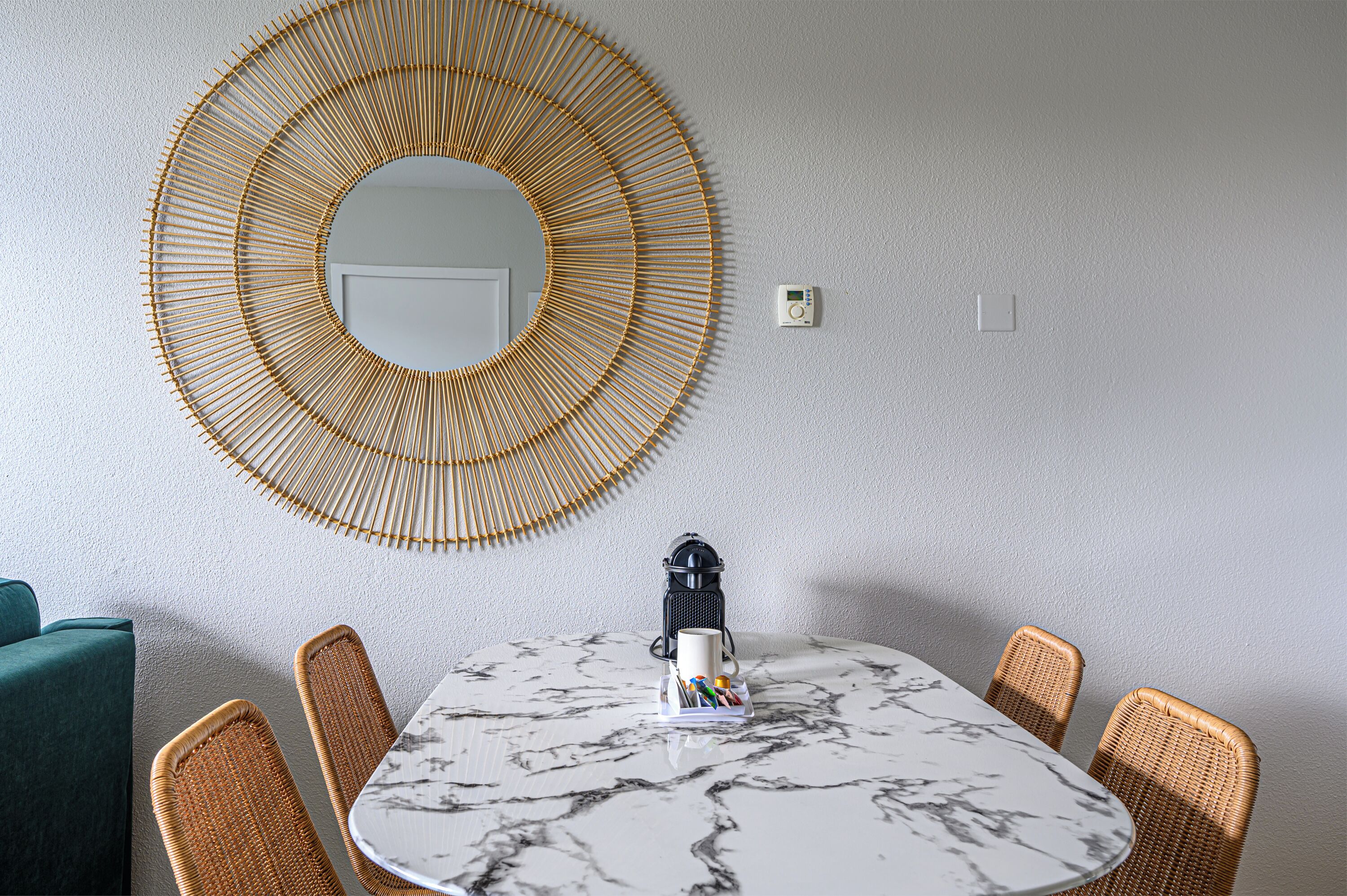 Un comedor con una mesa redonda, sillas de mimbre, una cafetera y un espejo colgado en la pared.