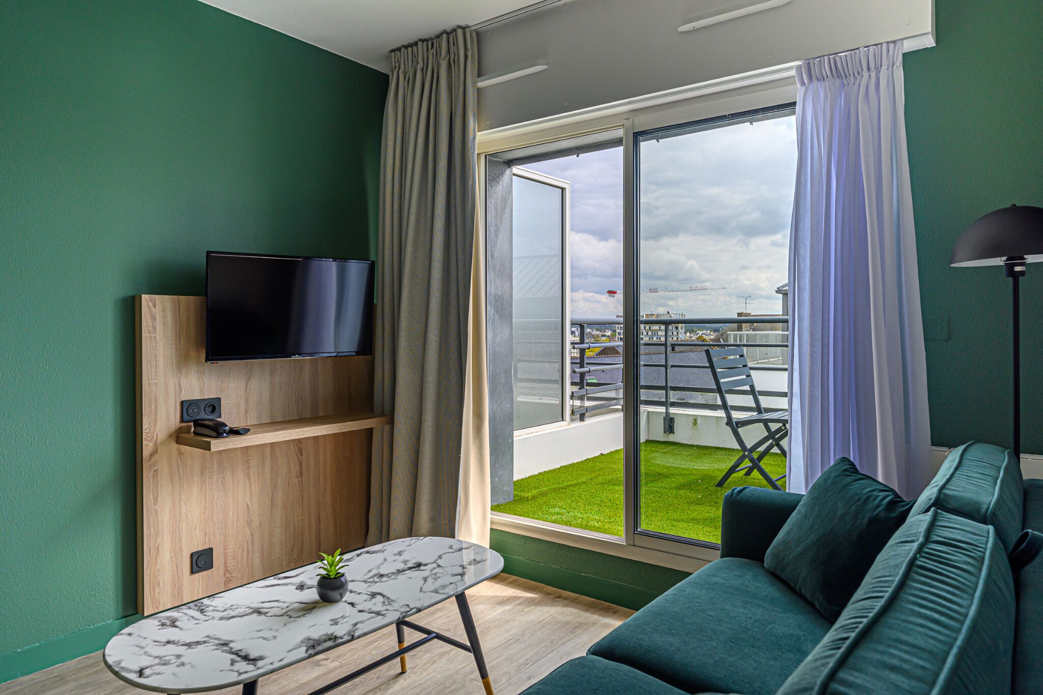Ett modernt vardagsrum med en platt-TV, en soffa, ett soffbord och en balkong med utsikt över staden.