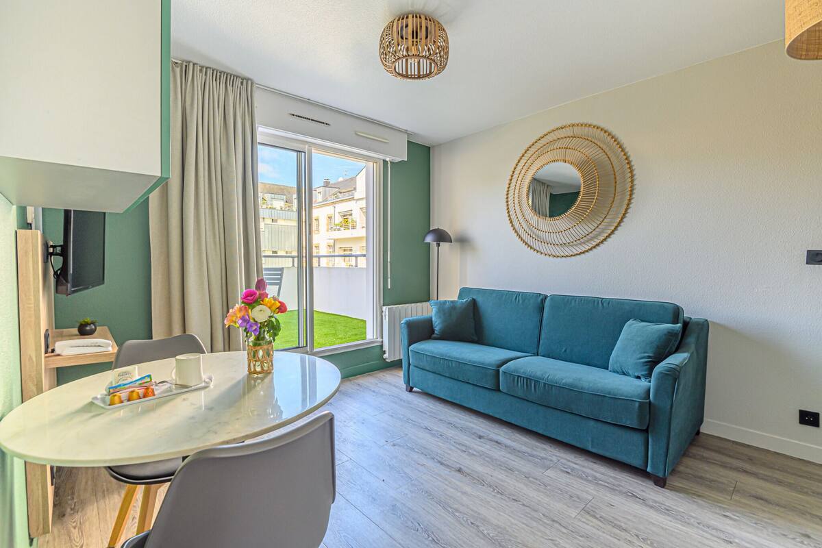 A modern living room with a teal sofa, a round coffee table, and a large window with a view of buildings outside.