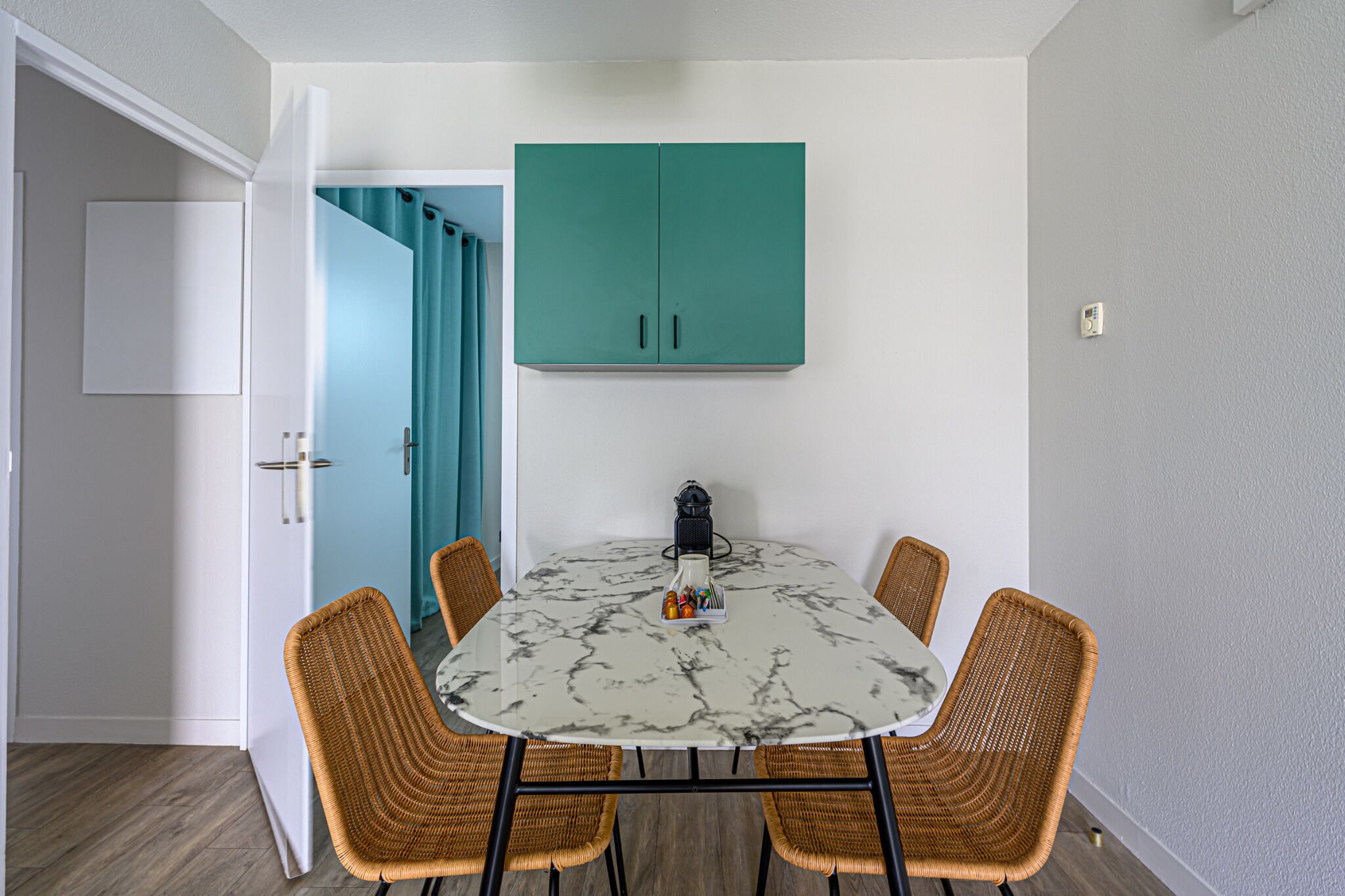 Un comedor con una mesa redonda, cuatro sillas de mimbre, un armario azul turquesa arriba y una puerta blanca con cortina azul.