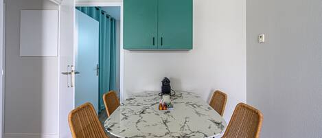 Un comedor con una mesa redonda, cuatro sillas de mimbre, un armario azul turquesa arriba y una puerta blanca con cortina azul.