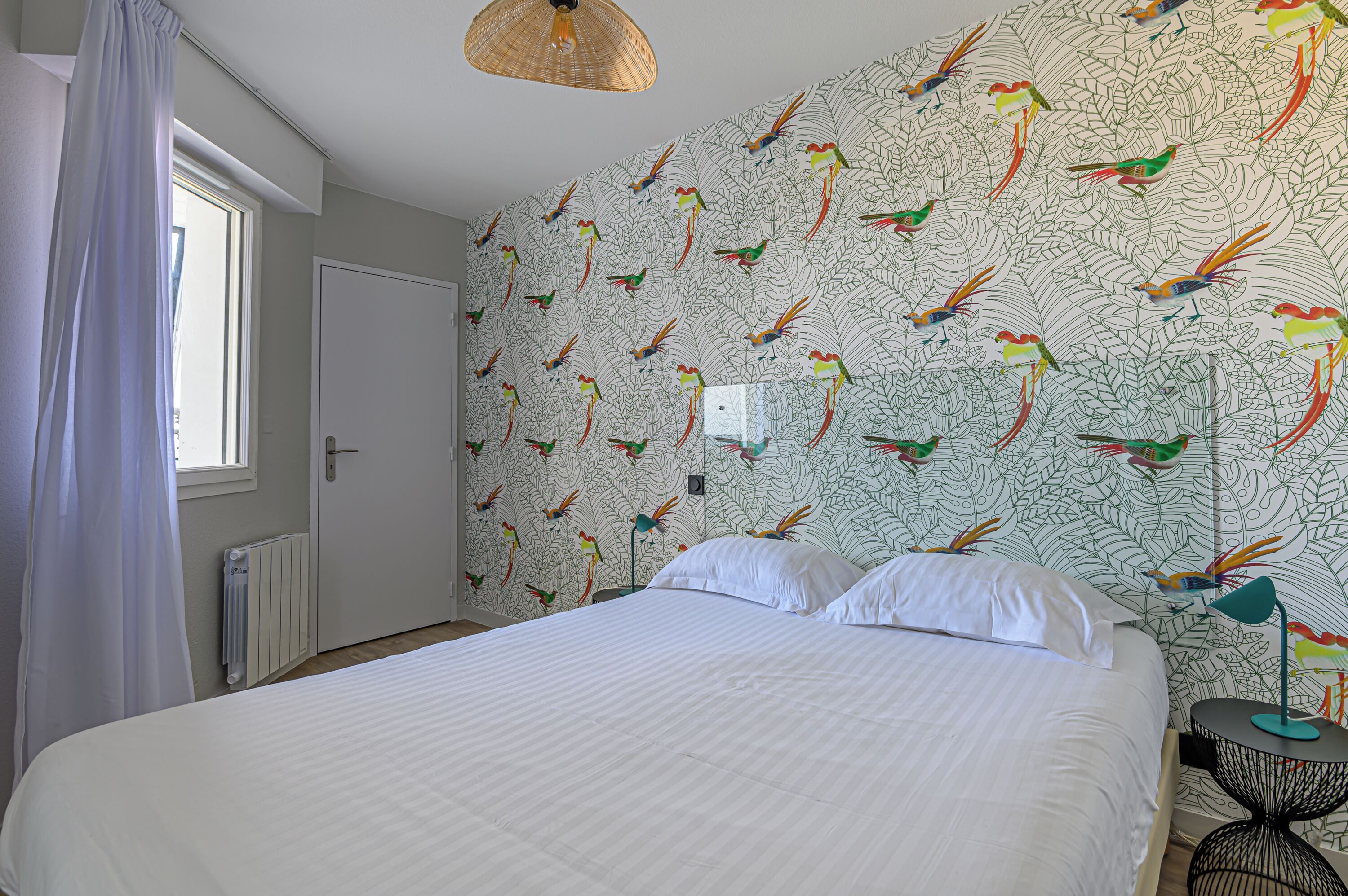 Une chambre avec un grand lit, du linge de lit blanc, une fenêtre avec un rideau blanc et un mur orné d’un motif d’oiseaux.