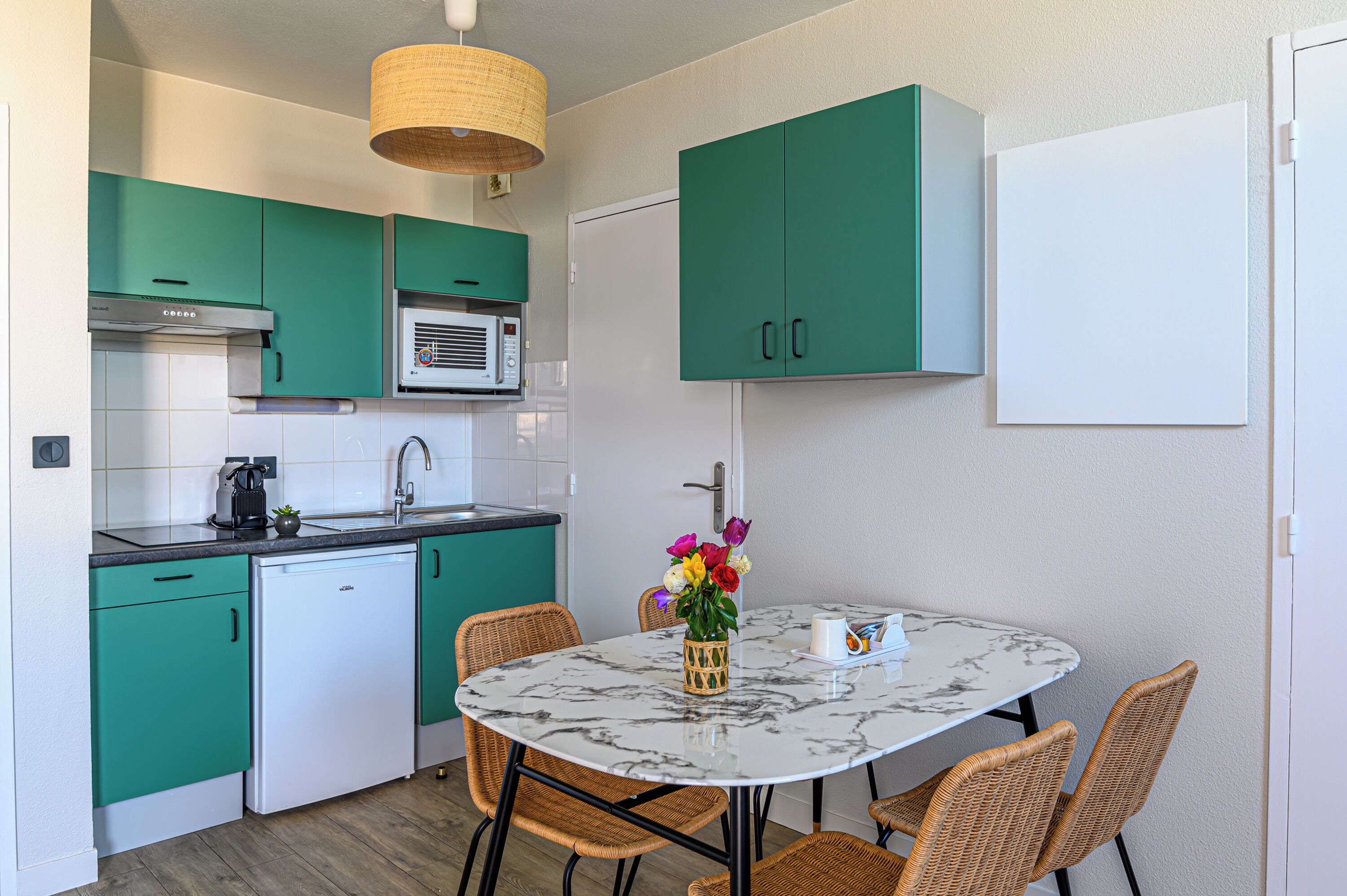 Una cocina compacta con armarios color turquesa, un refrigerador blanco y un microondas. Hay una mesa de comedor con sillas de mimbre y un jarrón con flores.