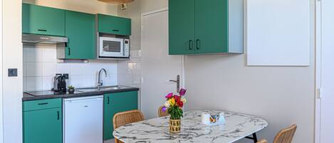 Una cocina compacta con armarios color turquesa, un refrigerador blanco y un microondas. Hay una mesa de comedor con sillas de mimbre y un jarrón con flores.