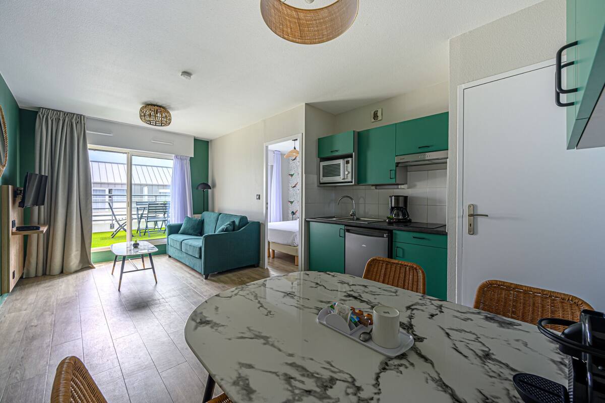 A modern living room with a sofa, a dining table with a marble top, and a kitchen area with teal cabinets and a microwave.