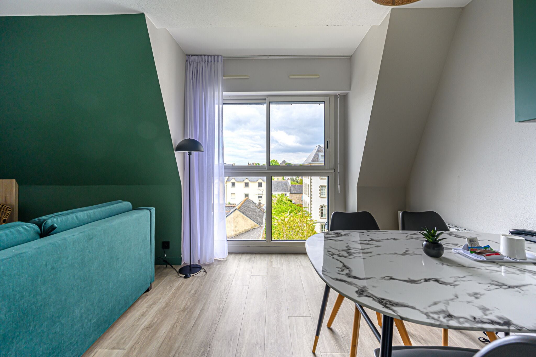 En moderne stue med en turkis sofa, et spisebord med stole, et stort vindue med gardiner og udsigt til bygninger.