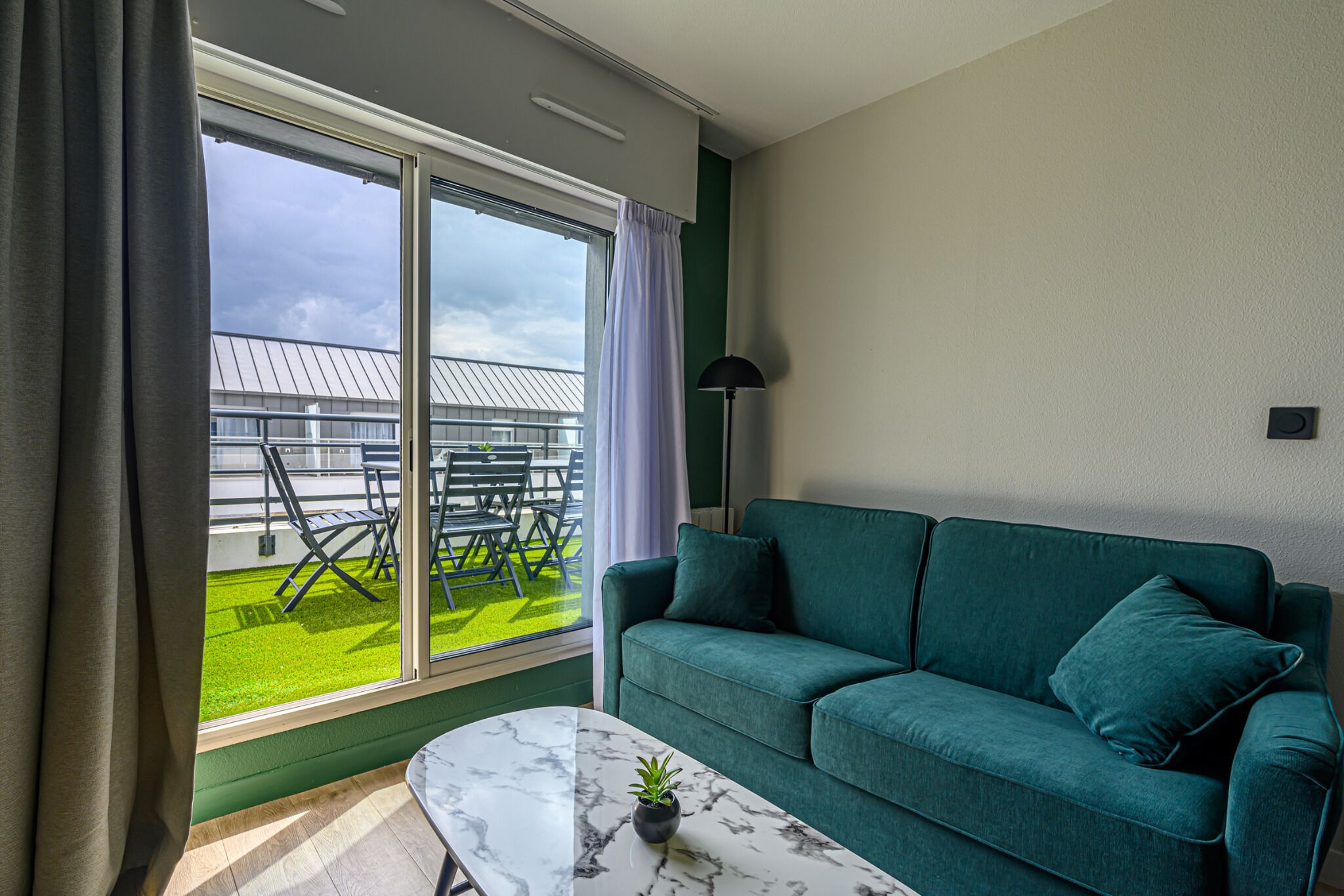 A modern living room with a green sofa, a marble coffee table, and a view of a balcony with outdoor furniture.