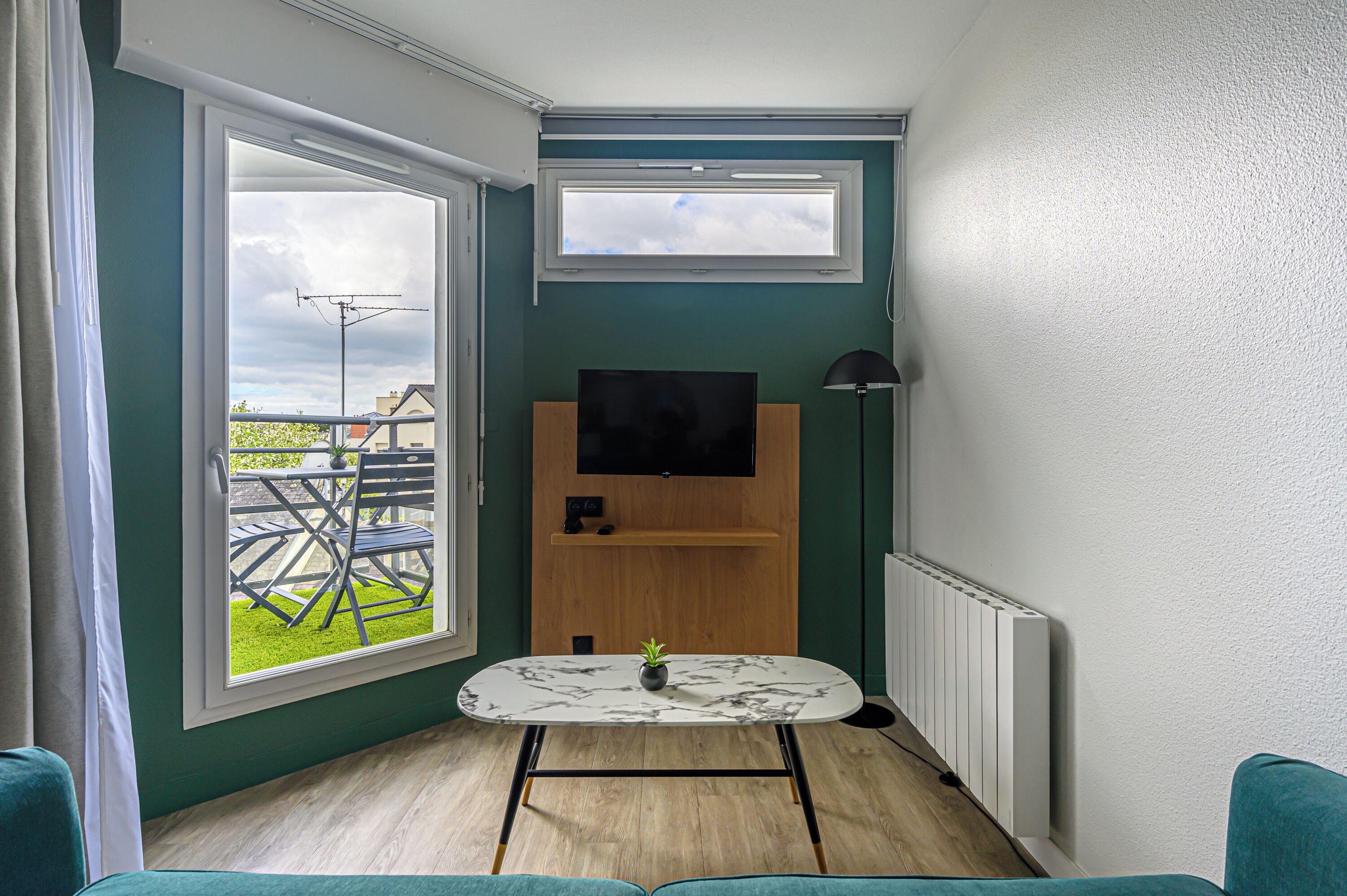 Ein kleiner, moderner Raum mit einem an der Wand befestigten Fernseher, einem Tisch mit einer Pflanze und einem Blick ins Freie durch ein großes Fenster.