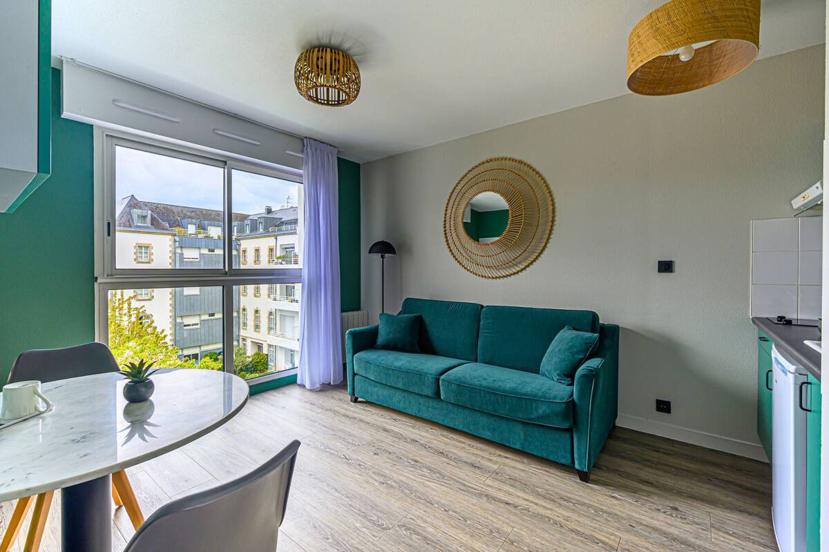 A modern living room with a green sofa, a round mirror, and a dining area with a table and chairs.