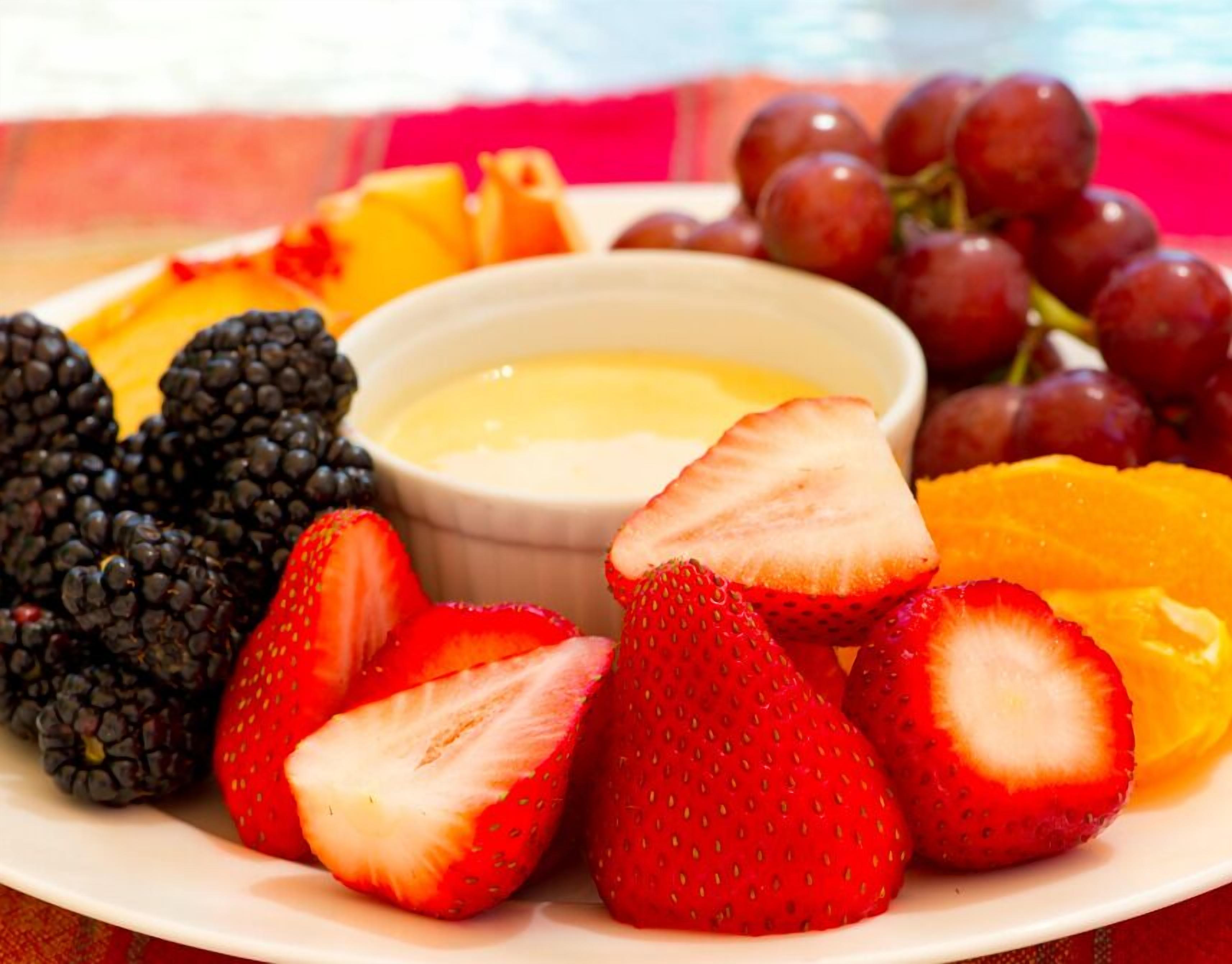 Plato con frutas variadas, incluyendo fresas, zarzamoras, uvas y una rodaja de naranja, acompañado de un recipiente pequeño con salsa.