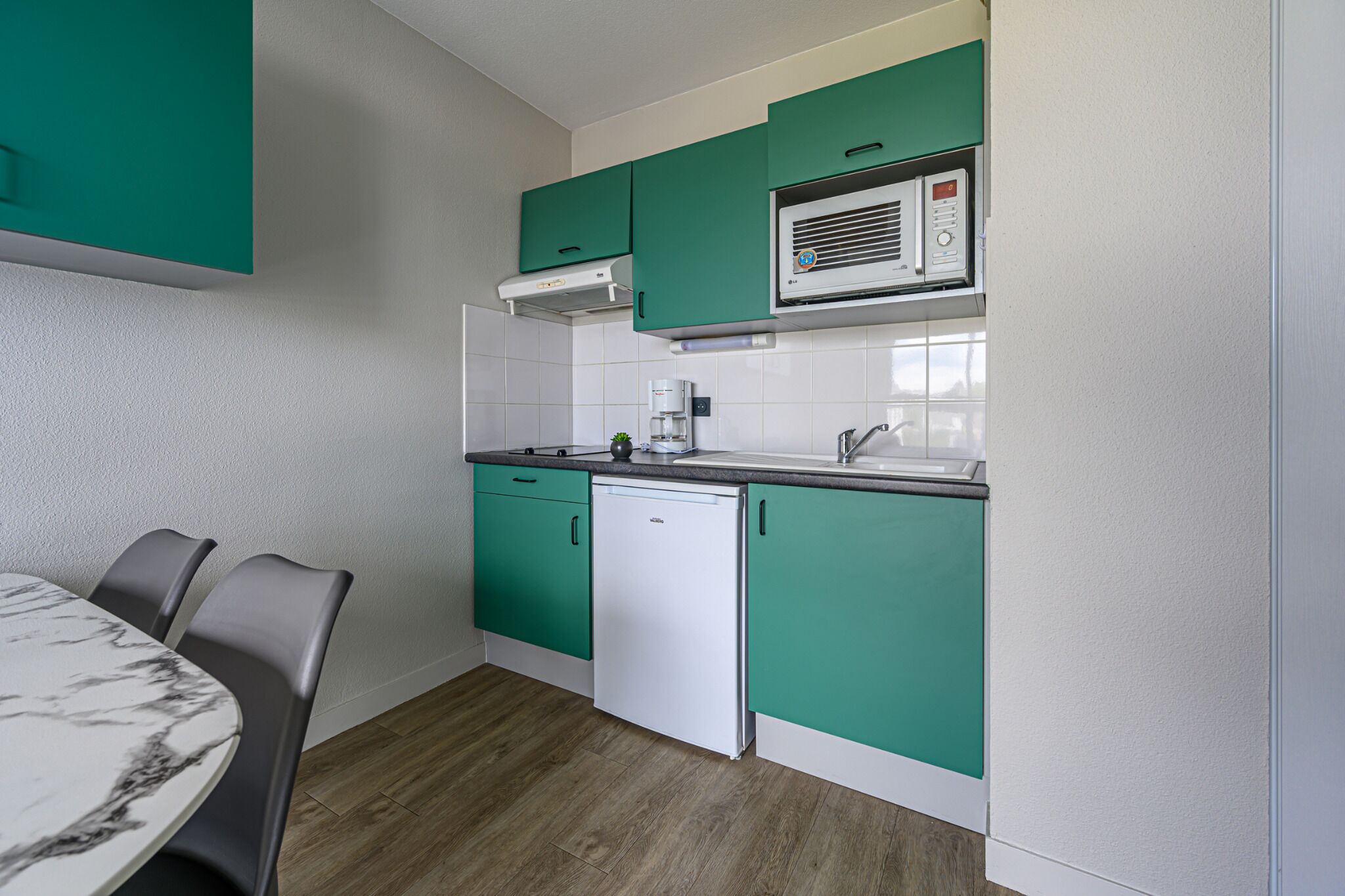 Een kleine keuken met blauwgroene kastjes, een witte vaatwasser en een eettafel met marmeren blad en stoelen.