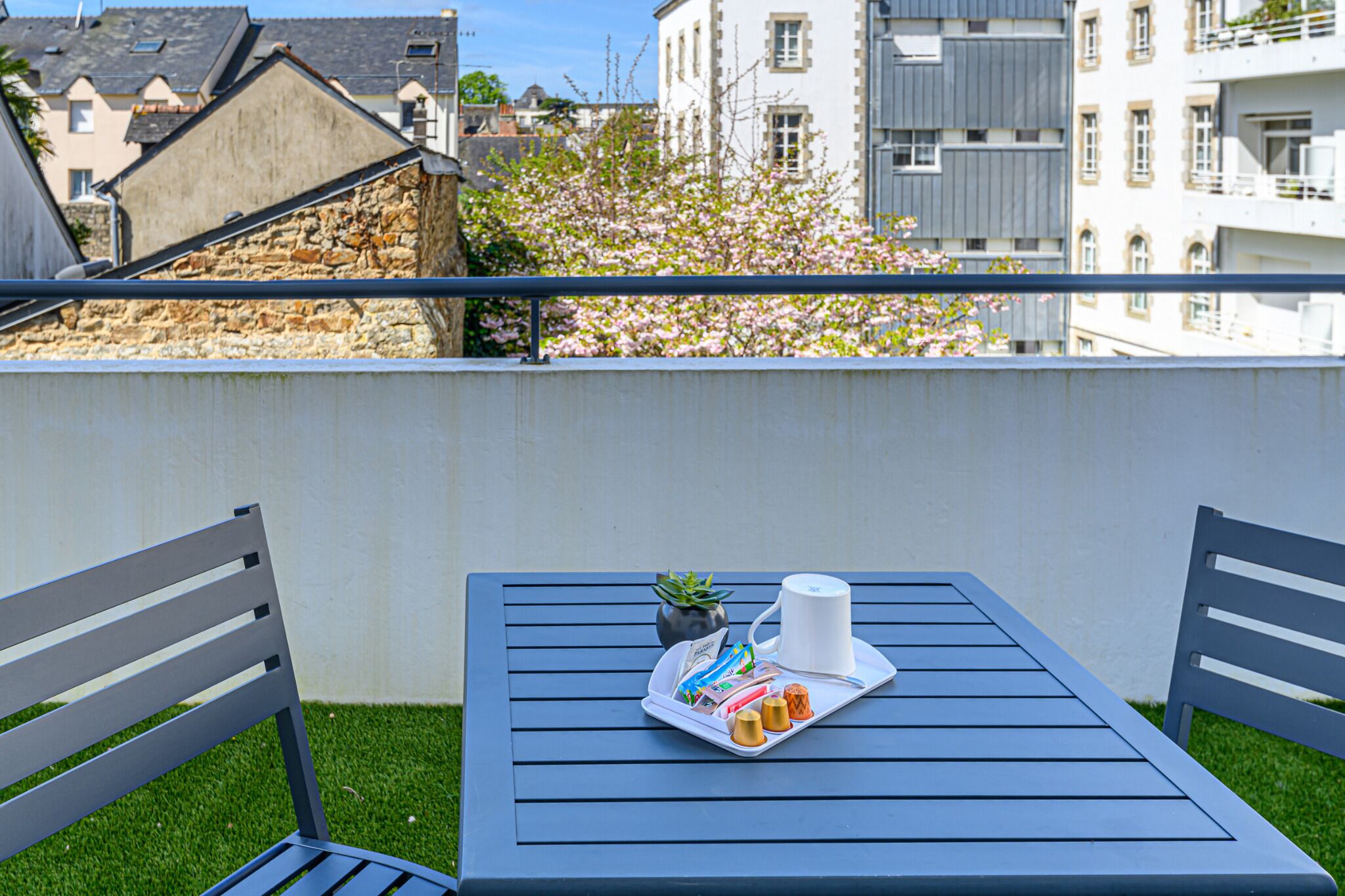 Un balcon avec une table et des chaises, un plateau avec de la nourriture et une tasse, et une vue sur des immeubles et un arbre.