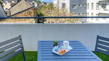 Un balcon avec une table et des chaises, un plateau avec de la nourriture et une tasse, et une vue sur des immeubles et un arbre.