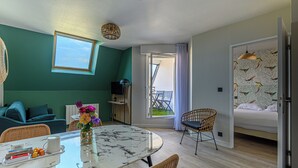 Una habitación moderna con una mesa de comedor, sillas de mimbre, un sofá, una cama con cabecera estampada y un balcón con vistas.