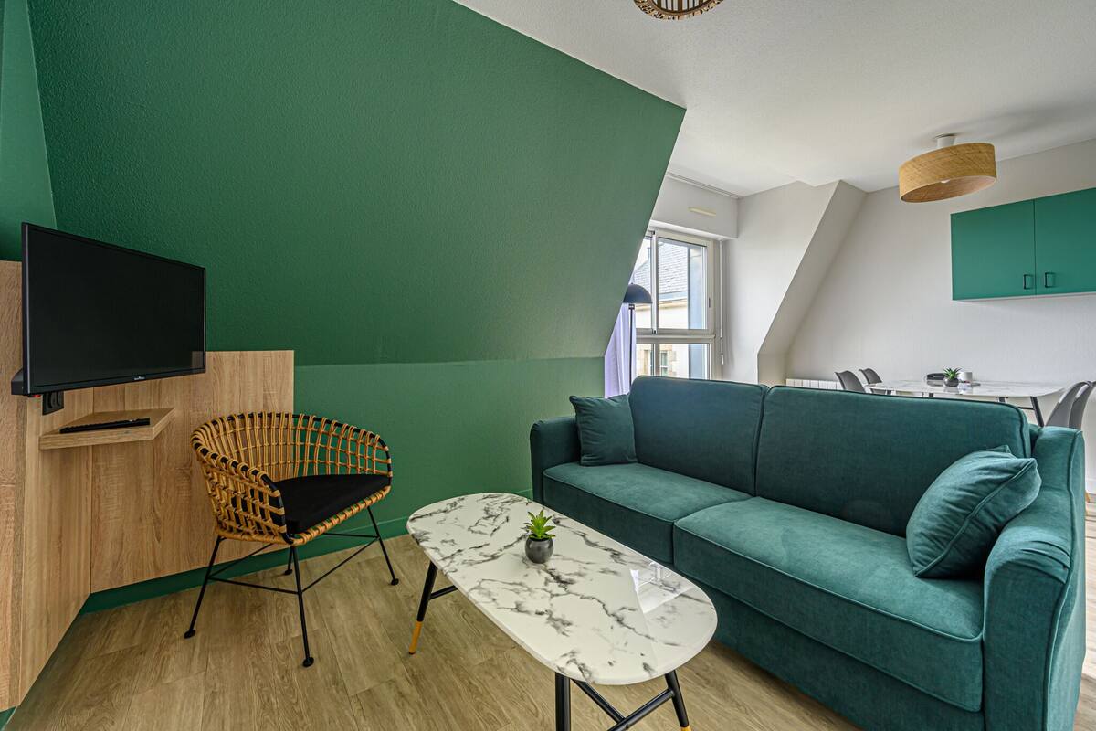 A modern living room with a green wall, a sofa, a coffee table, a TV, and a wicker chair.