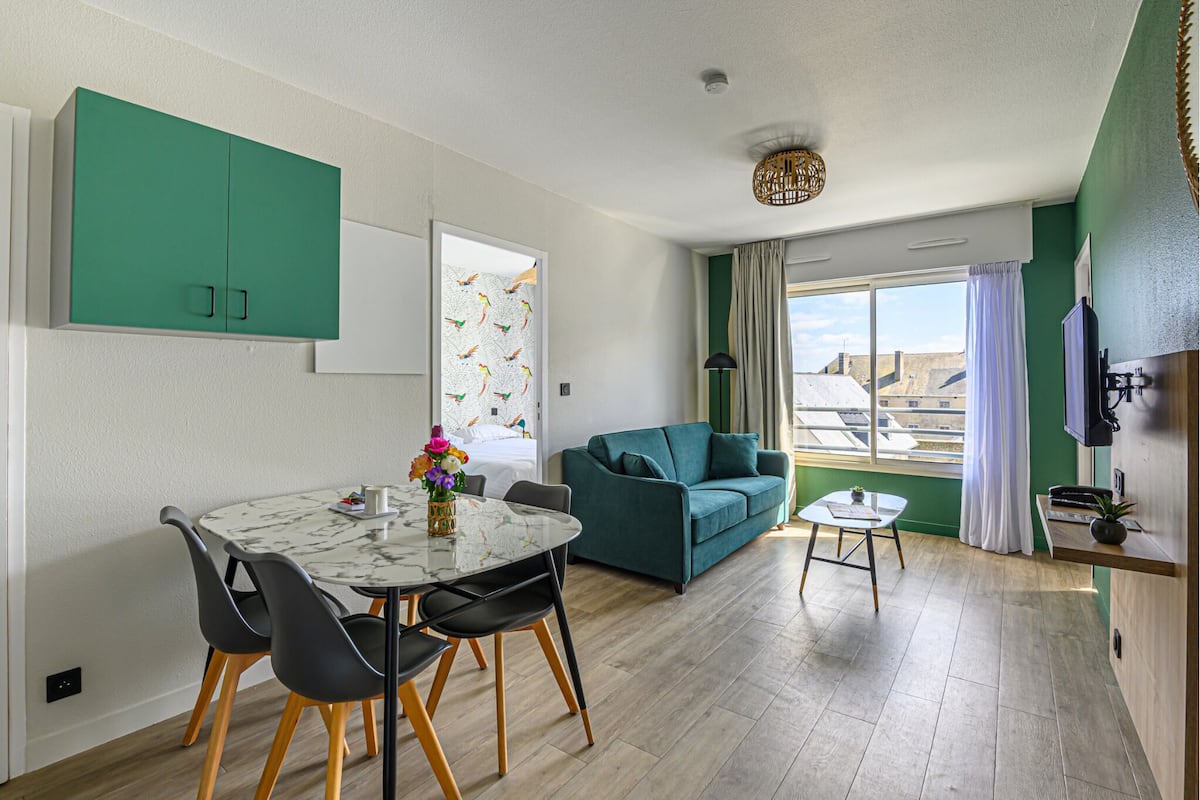A modern living room with a teal sofa, a dining table with chairs, and a small side table.