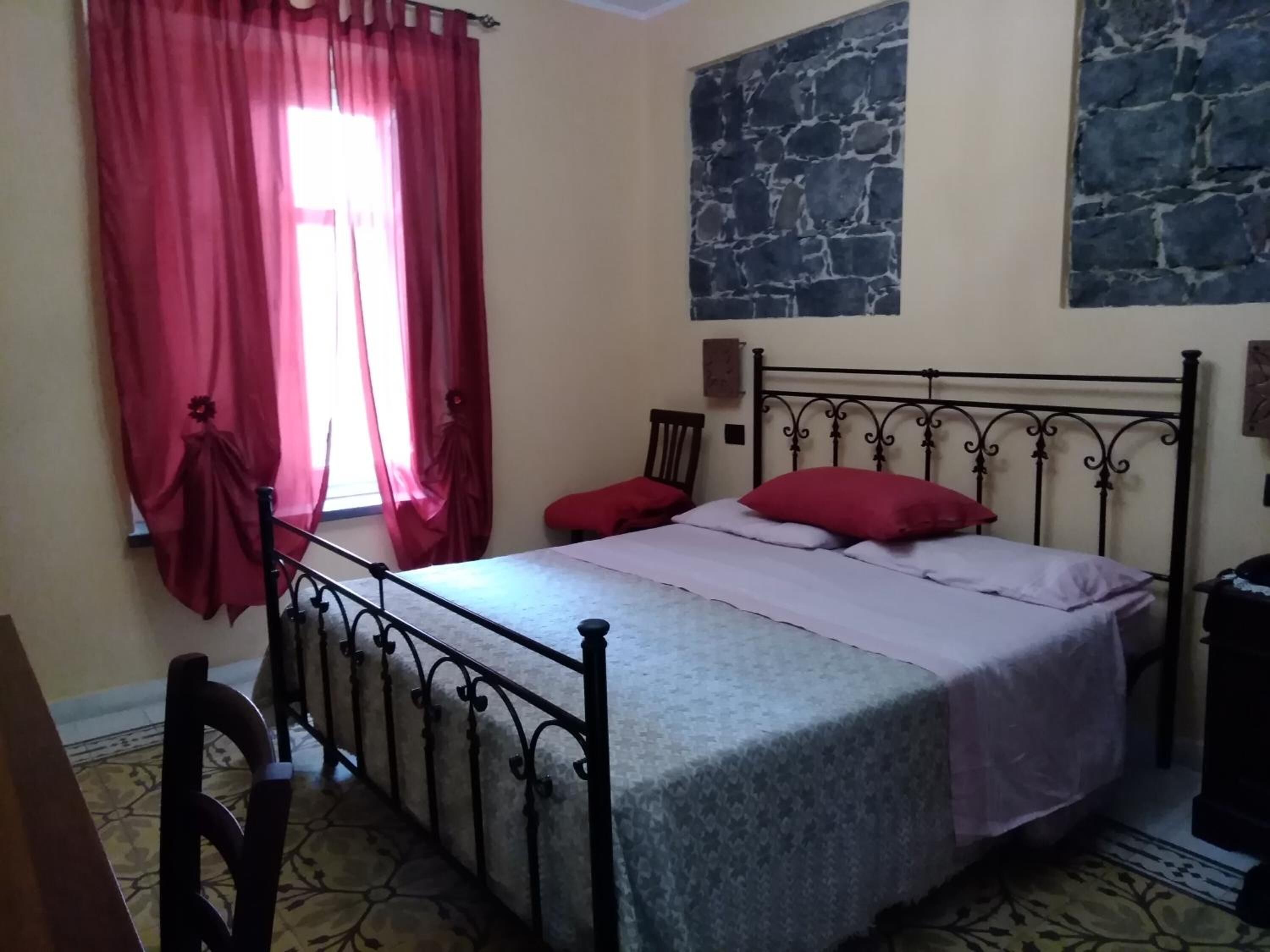Una camera da letto con un letto, una sedia, una finestra con tende rosse e un muro in pietra.