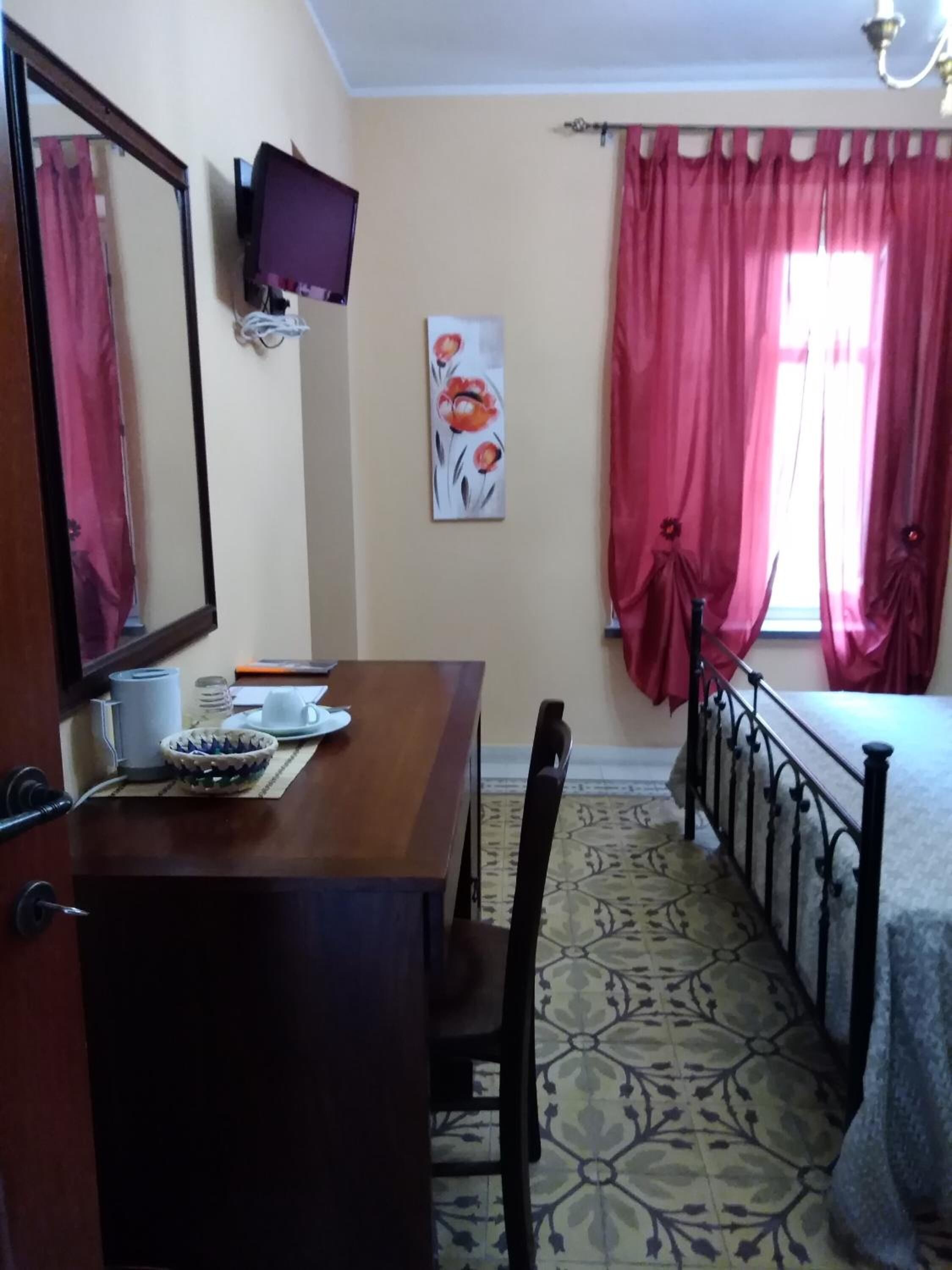 Una habitación con un escritorio de madera, una silla, una cama con estructura metálica, un televisor montado en la pared y una ventana con cortinas rojas.