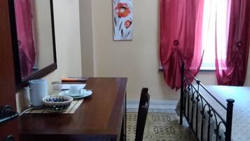 Una habitación con un escritorio de madera, una silla, una cama con estructura metálica, un televisor montado en la pared y una ventana con cortinas rojas.