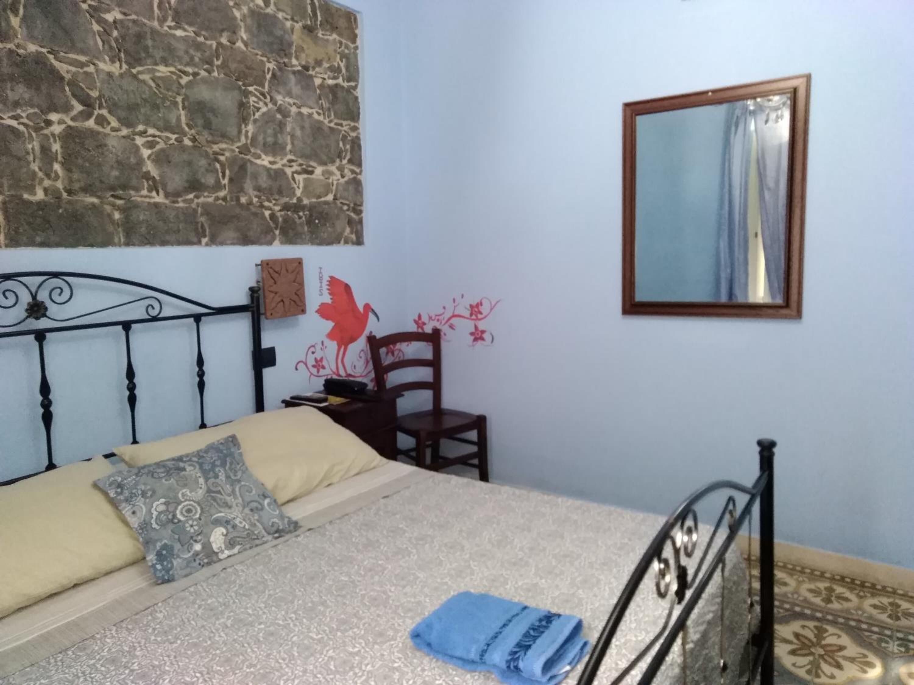 A bedroom with a stone wall, a bed with patterned pillows, a wooden chair, a small table, and a mirror on the wall.