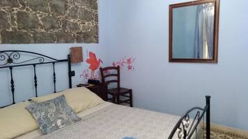 A bedroom with a stone wall, a bed with patterned pillows, a wooden chair, a small table, and a mirror on the wall.