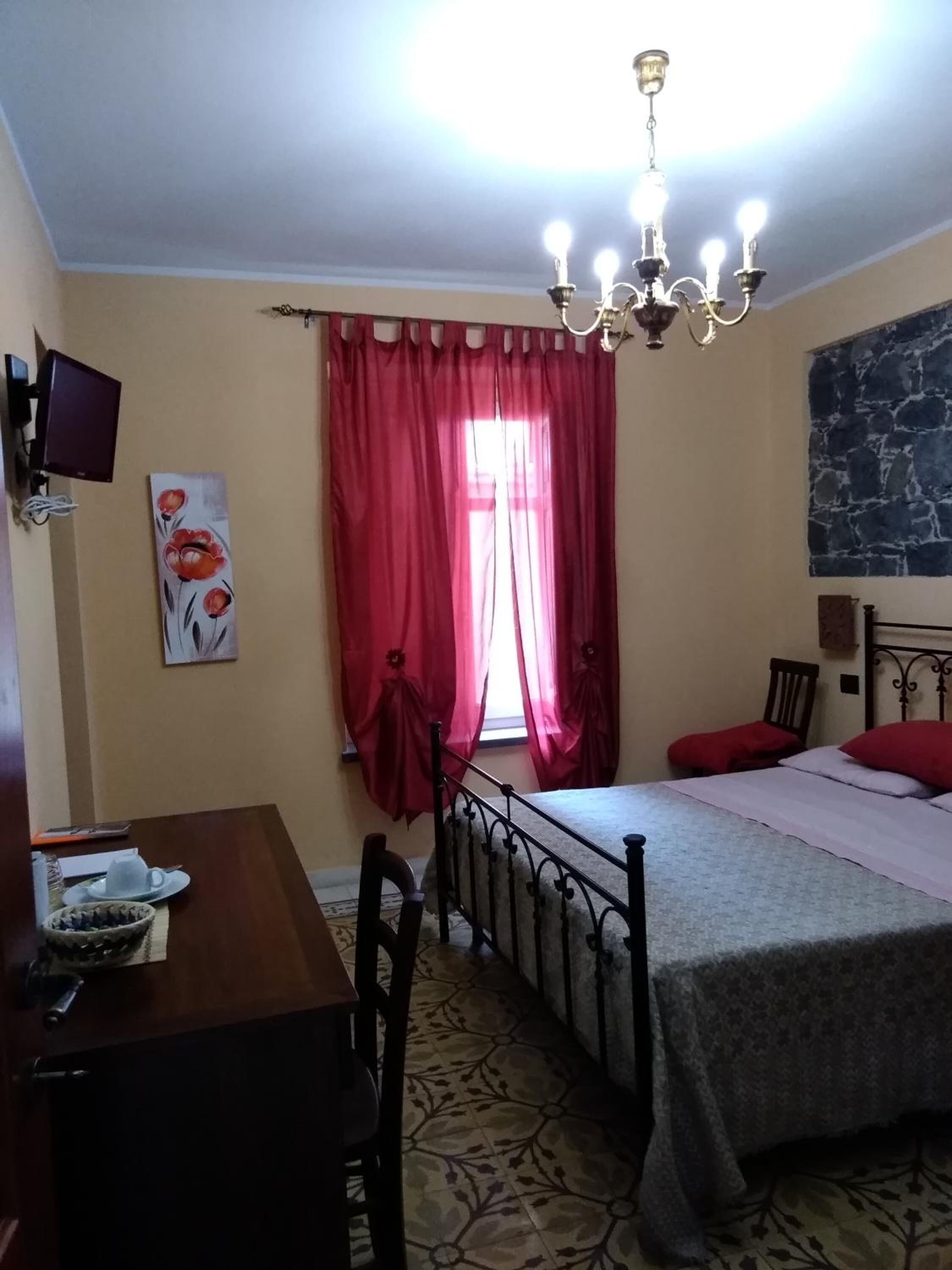 Un dormitorio con cama, escritorio, silla, televisor y ventana con cortinas rojas.