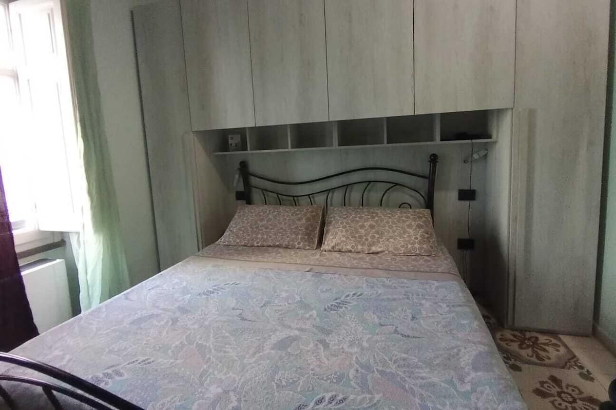 Ein Einzelbett mit einem gemusterten Bettbezug, ein Kopfteil mit Ablagefächern und ein Fenster mit Vorhängen.