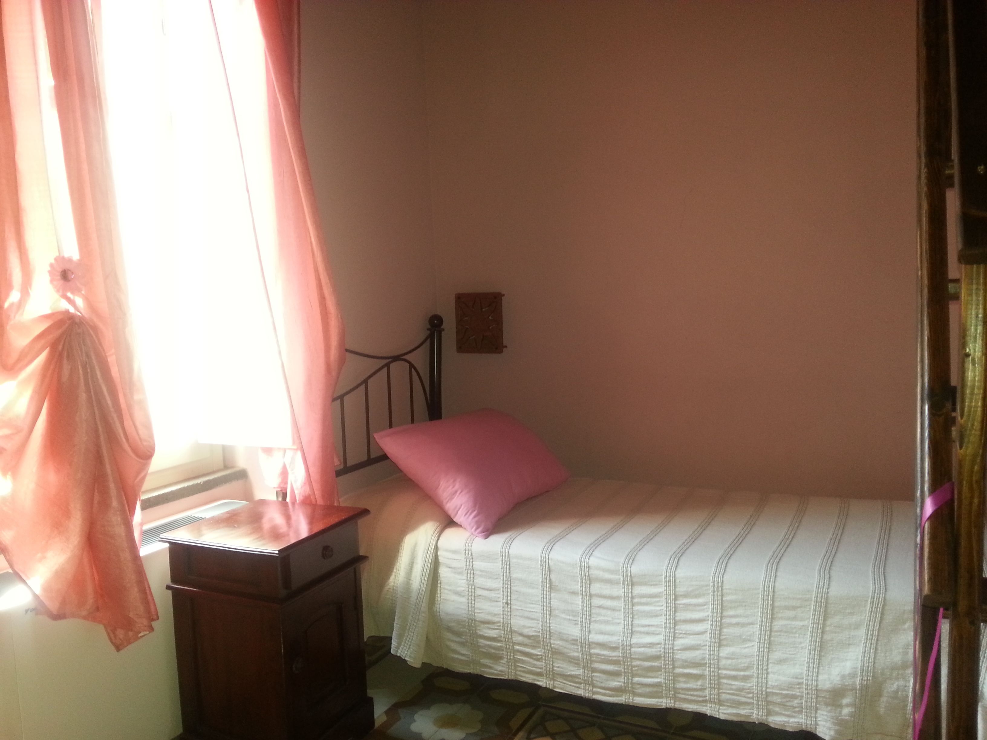 Un dormitorio con una cama individual, una mesita de noche, un ventanal con cortinas traslúcidas y un pequeño reloj de pared.