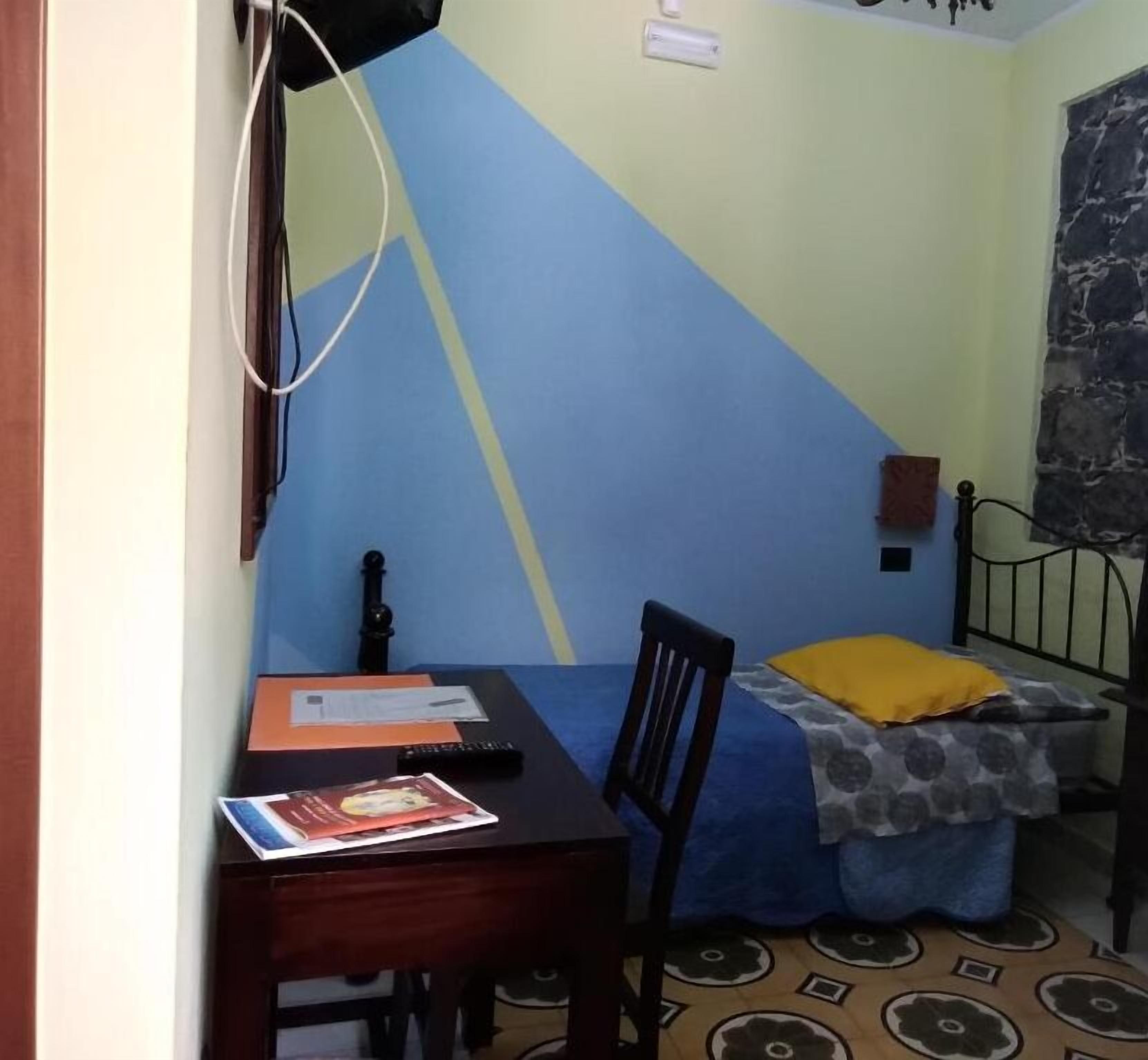 Une petite pièce comprenant un lit, un bureau et une chaise. On y trouve une fenêtre avec un rideau, une horloge murale et une prise de courant.