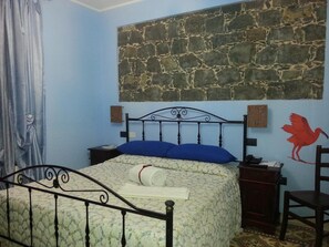 Un dormitorio con pared de piedra, una cama con colcha estampada, almohada azul, mesita de noche de madera, una silla y un mural en la pared con la imagen de un pájaro.