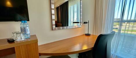 Una habitación de hotel con un escritorio de madera, una silla de oficina negra, un espejo grande, un televisor de pantalla plana y un ventanal con cortinas.