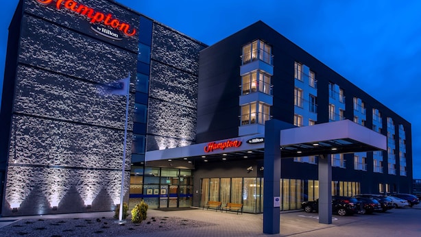 Un hôtel moderne, reconnaissable à son grand panneau « Hampton by Hilton », à ses larges fenêtres et à son entrée abritée.
