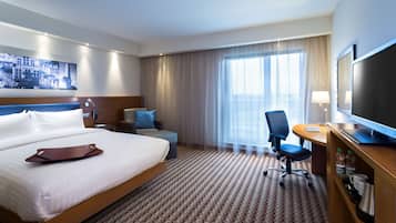 Ein Hotelzimmer mit einem großen Bett, einem Schreibtisch mit Fernseher, einem Sessel und einem fenster mit Vorhängen.